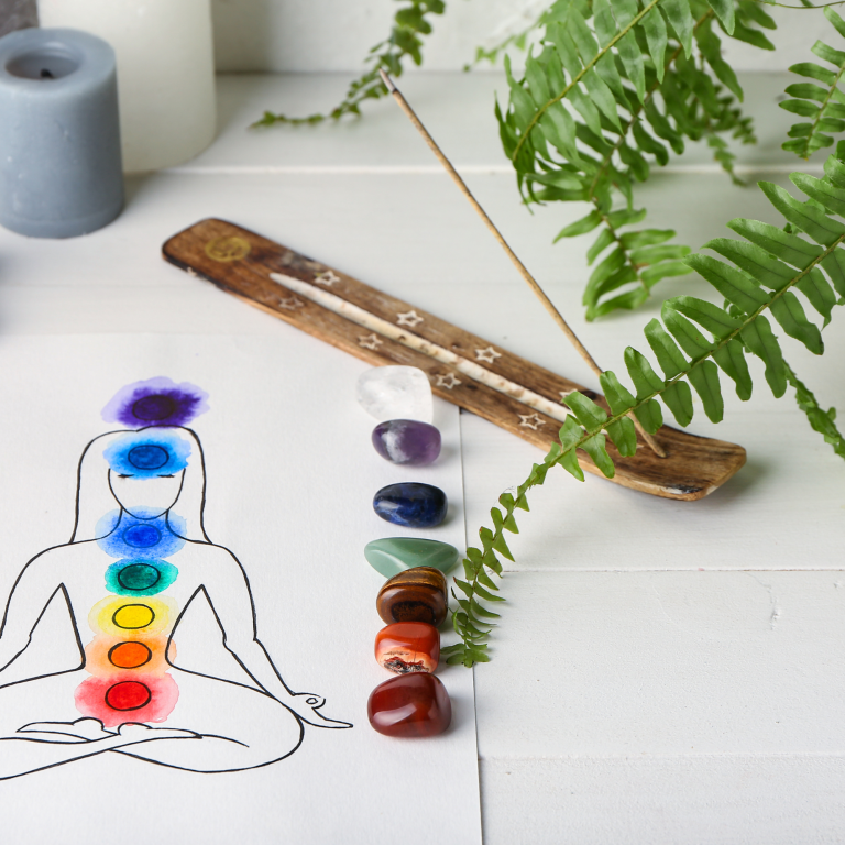 Chakra diagram with crystals, incense stick, candle, and a fern on a table.