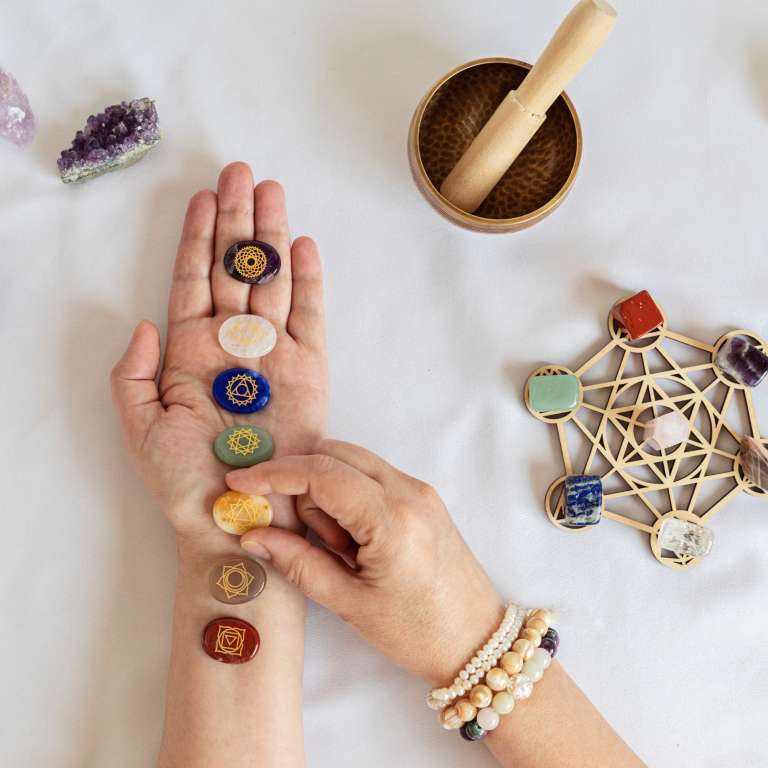 Hand with seven colourful crystals, surrounded by a bowl and geometric design on a white surface.